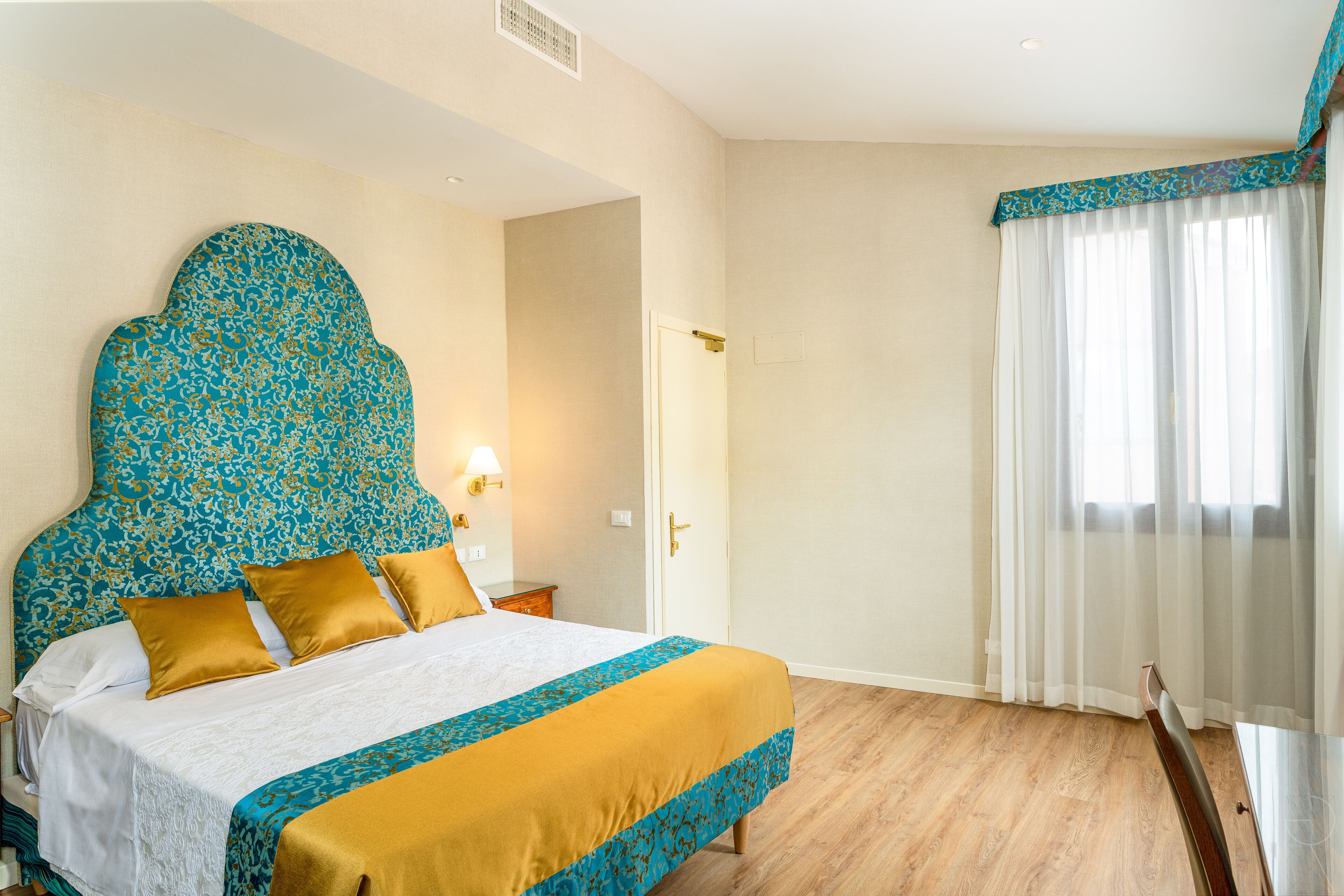 Ein Hotelzimmer mit einem großen Bett, Holzboden, einer Tür und einem Fenster mit transparenten Vorhängen.