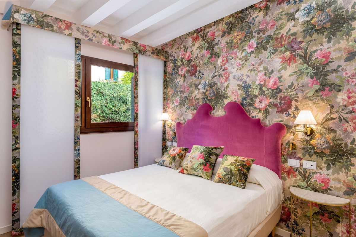 A bedroom with a floral wallpaper, a pink headboard, and a bedside table with a lamp.