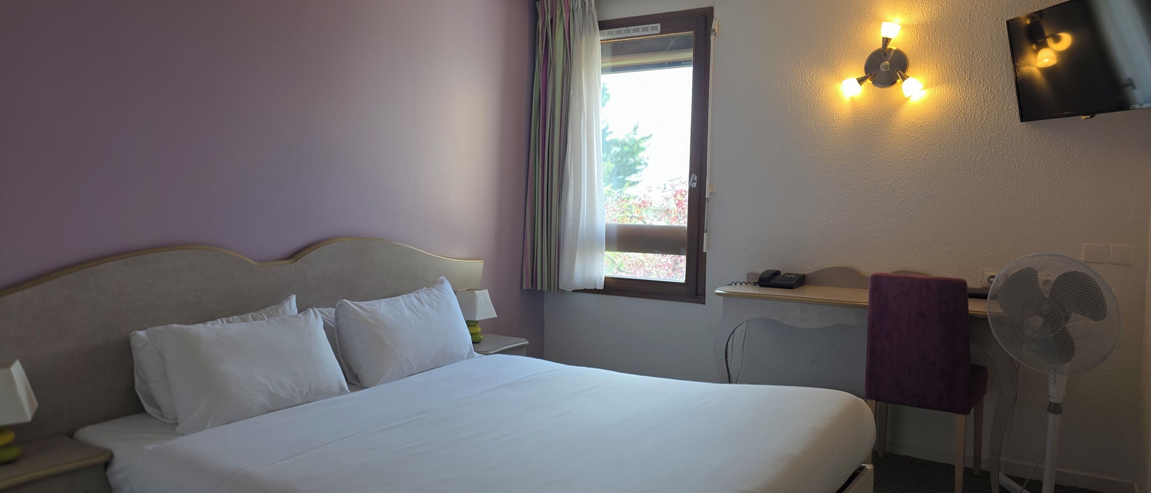 Une chambre d’hôtel comprenant un lit, un bureau, une chaise, une fenêtre, un ventilateur fixé au mur et un luminaire au plafond.