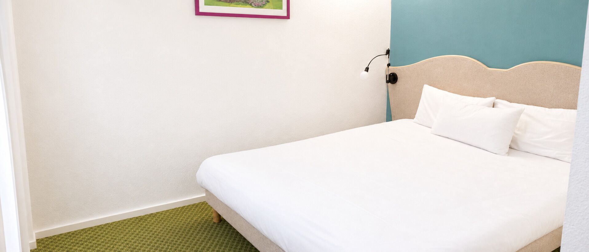 Una cama bien hecha con ropa de cama blanca, un tapete verde con textura, un cuadro colgado en la pared y un cabecero con una lámpara.
