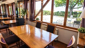 Un restaurante con mesas y sillas de madera, amplias ventanas con cortinas y plantas en macetas.