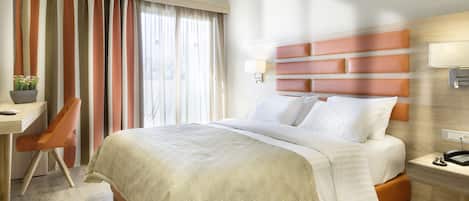 A hotel room with a large bed, a desk with a chair, a bedside table with a lamp, and a window with curtains.