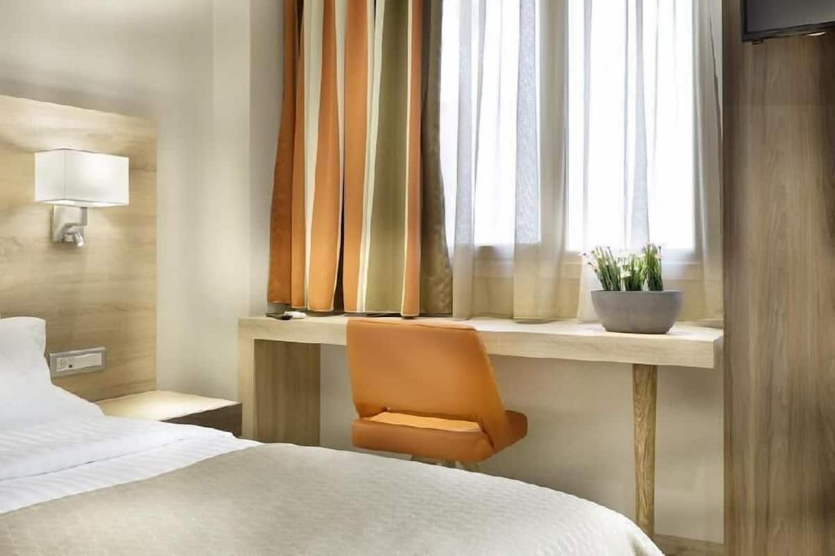 Habitació d'hotel amb llit, escriptori amb cadira, planta en test, làmpada penjada a la paret i finestra amb cortines.