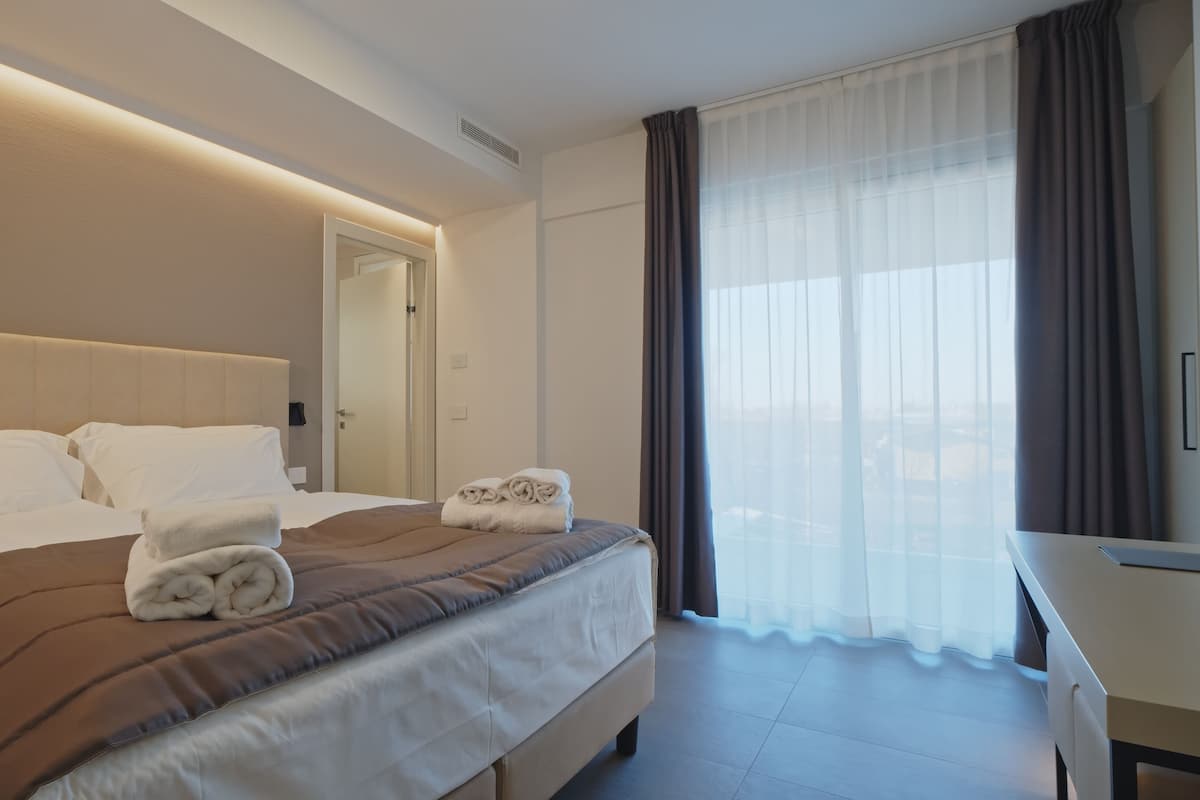 A hotel room with a large bed, a desk, and a view through sheer curtains.