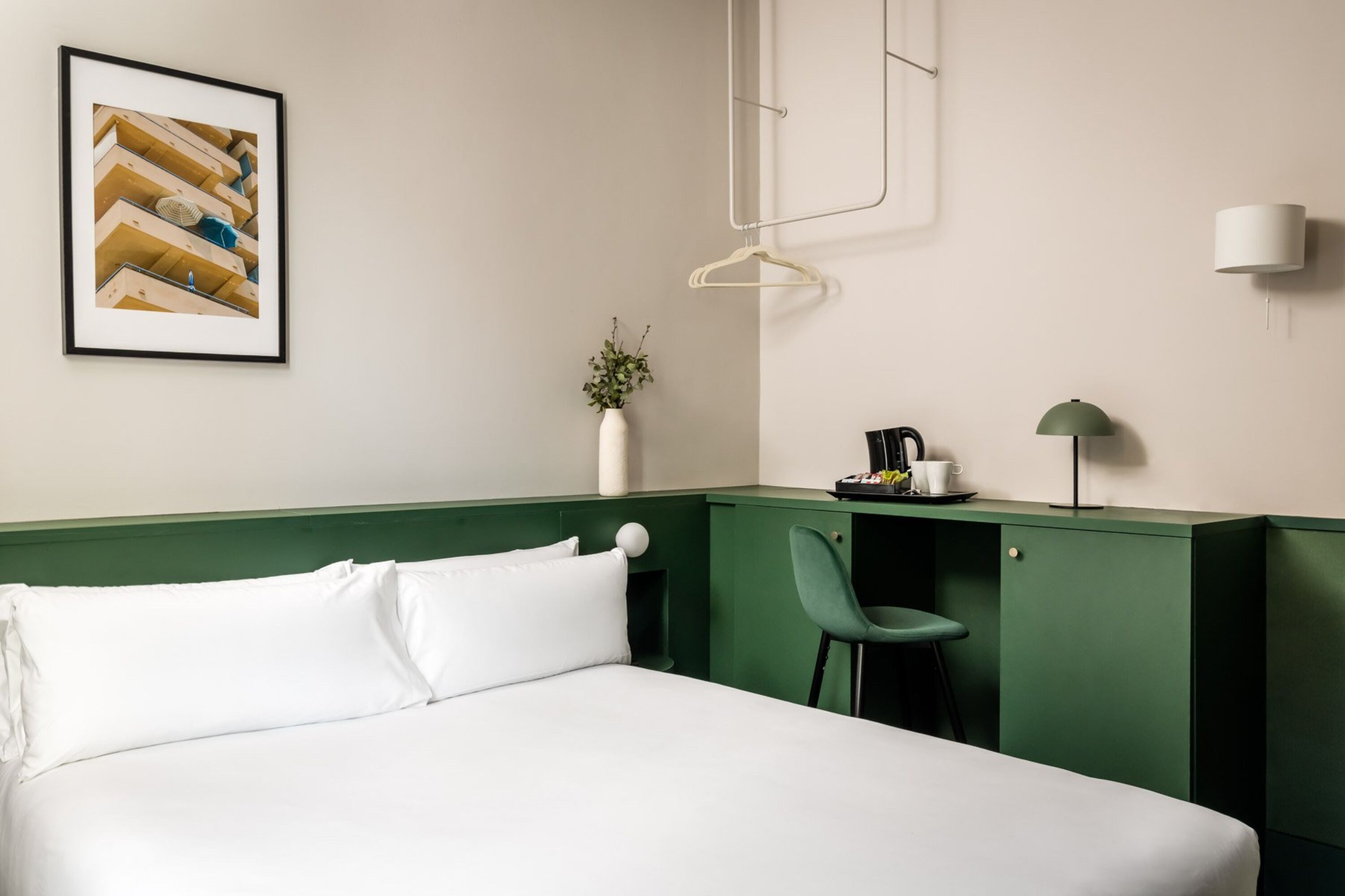 Une chambre d’hôtel comprenant un lit, un bureau avec une chaise, une lampe suspendue, un tableau encadré et un vase avec une plante.