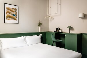 Quarto de hotel com cama, escrivaninha com cadeira, luminária pendente, quadro emoldurado e vaso com planta.