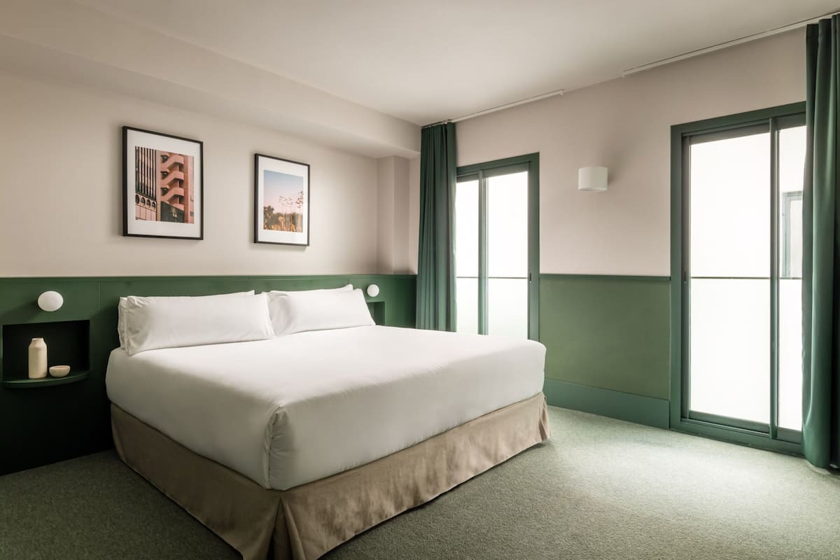 A neatly made bed in a hotel room with a green accent wall, a large window with curtains, and two framed pictures on the wall.