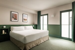 Una cama bien hecha en una habitación de hotel con una pared verde decorativa, un ventanal con cortinas y dos cuadros en la pared.