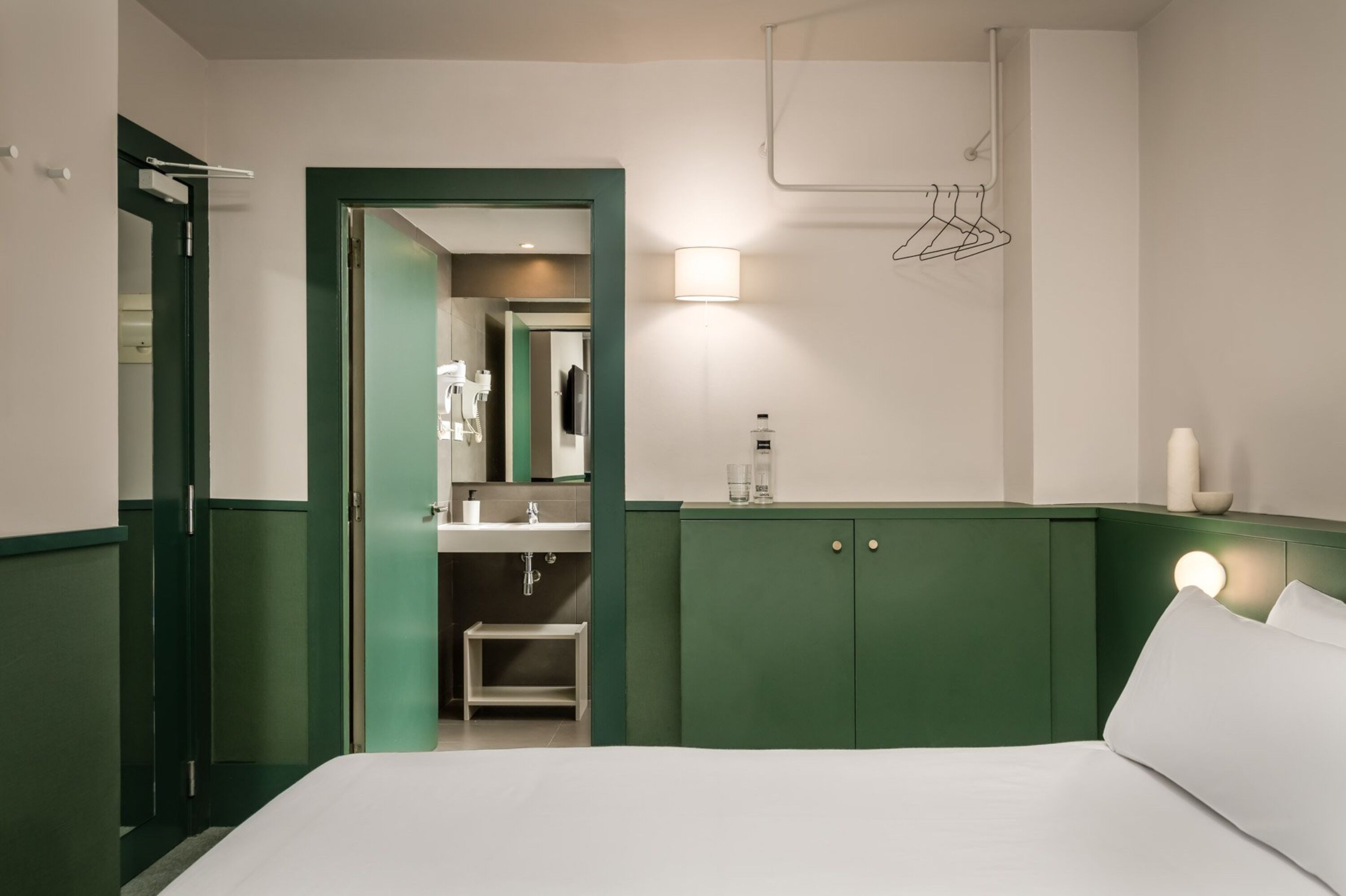 A hotel room with a bed, a green-framed door, a bathroom with a sink and mirror, and a wall-mounted towel rack.