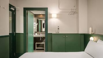 A hotel room with a bed, a green-framed door, a bathroom with a sink and mirror, and a wall-mounted towel rack.