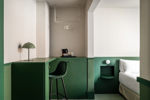 Una habitación de hotel compacta con una pared verde de acento, un escritorio pequeño con una silla, una cama y una mesita de noche con lámpara.