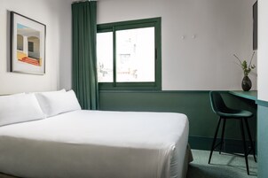 Habitación de hotel con cama, escritorio, silla, ventana con cortinas e imagen en un marco en la pared.