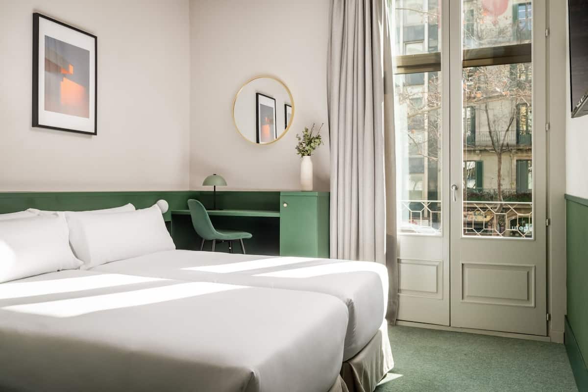 A hotel room with a large bed, a desk, a chair, a mirror, and a view of the outside.