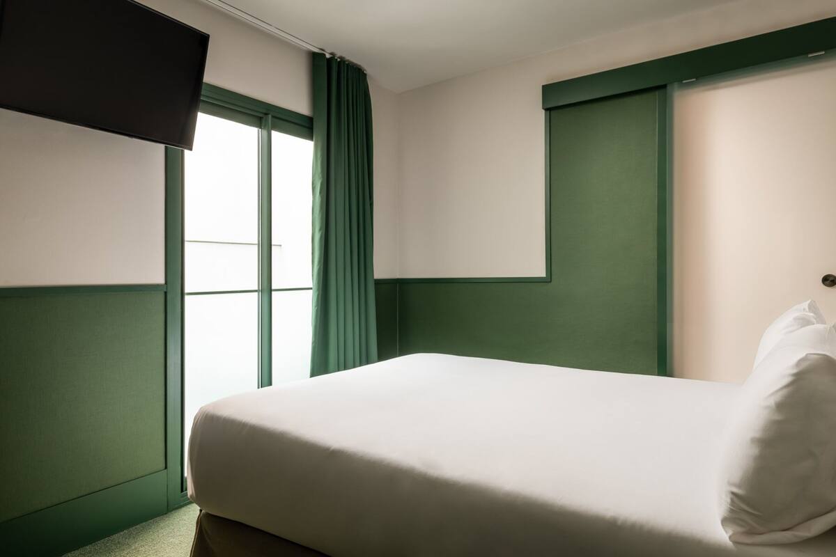 A hotel room with a bed, a flat-screen TV mounted on the wall, and a window with green curtains.