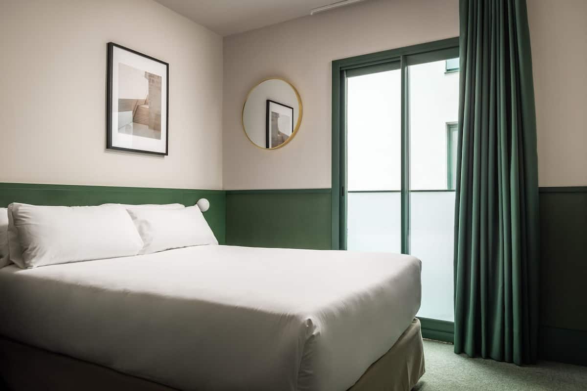A hotel room with a bed, a framed picture on the wall, a round mirror, and a window with curtains.