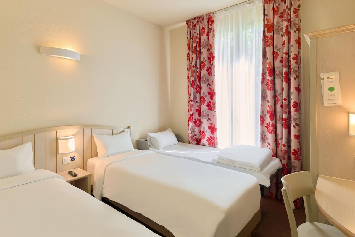 A hotel room with two beds, a bedside table with a lamp, a chair, and a window with floral curtains.