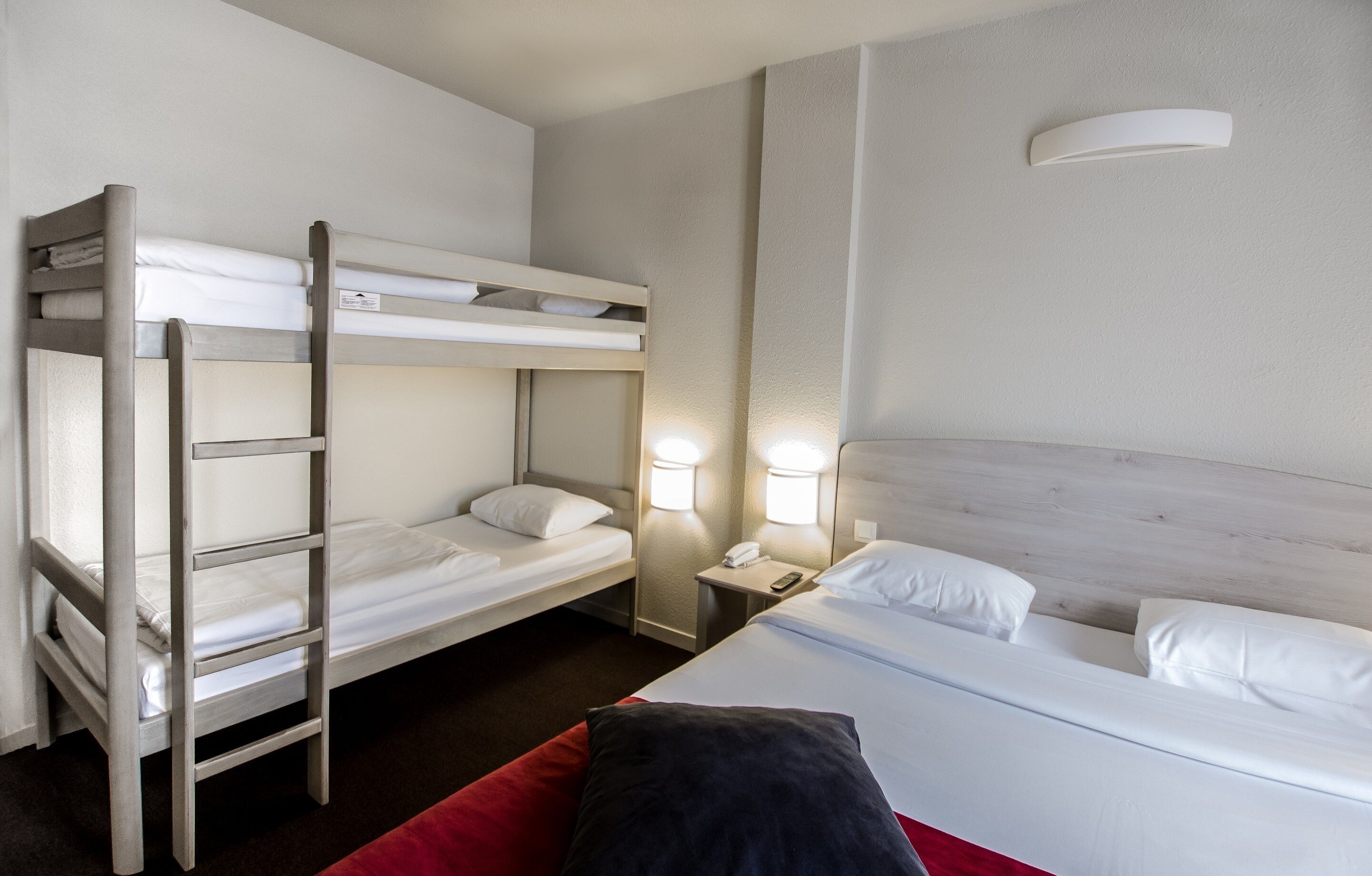 Habitación de hotel con literas, una cama individual, una mesita de noche con lámpara y un aplique de pared.