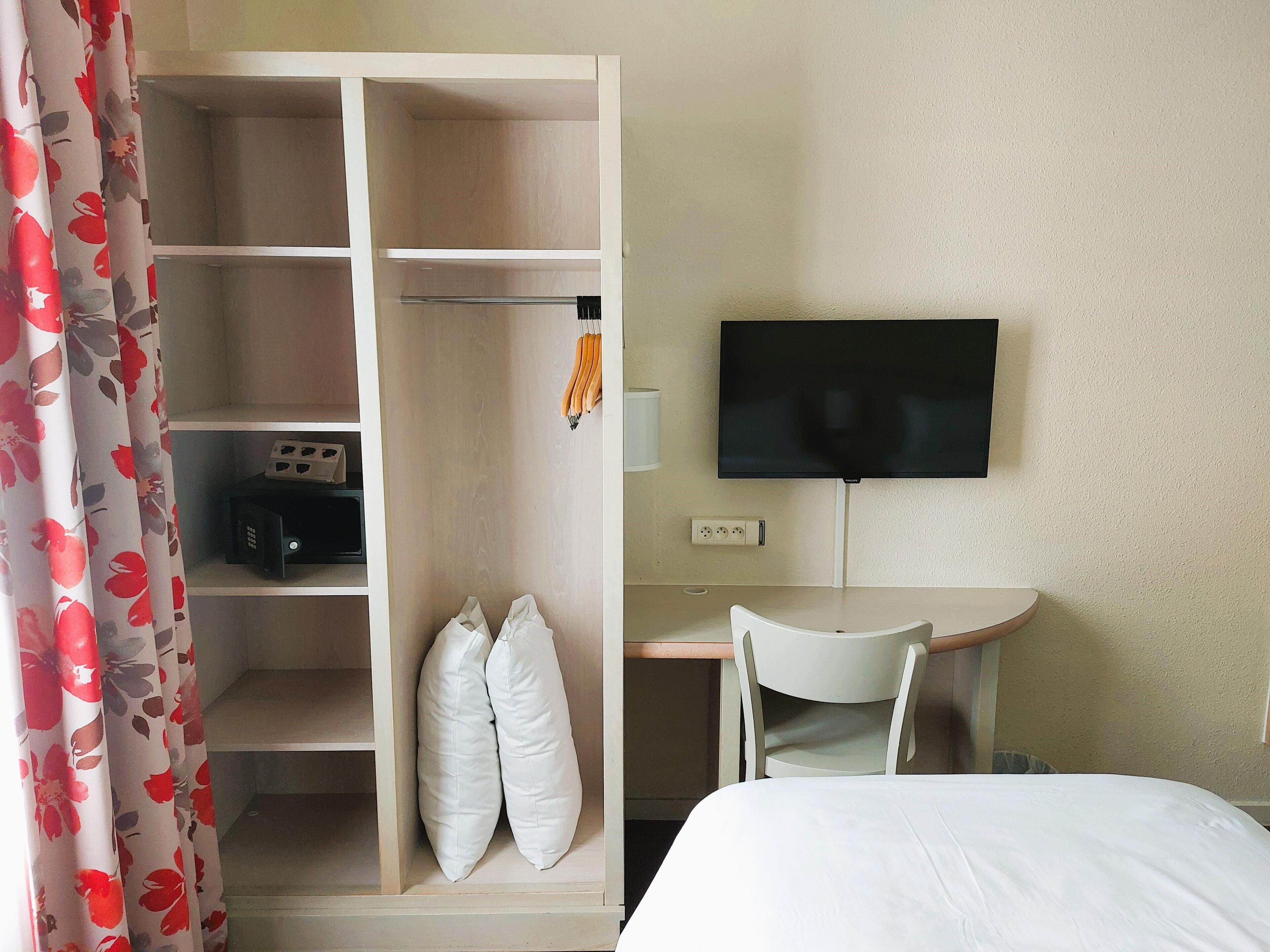A hotel room with a wardrobe, a desk with a chair, a bed, and a television.