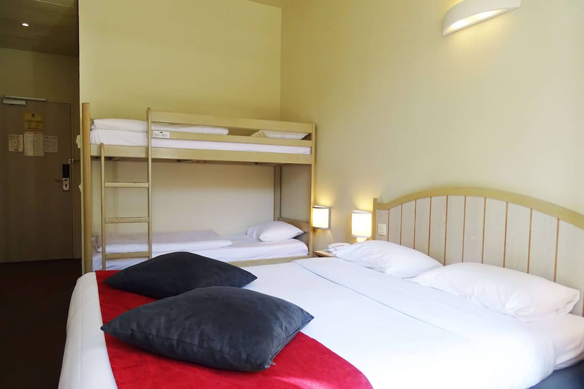 A hotel room with a bunk bed, a single bed with a red blanket, and two bedside lamps.