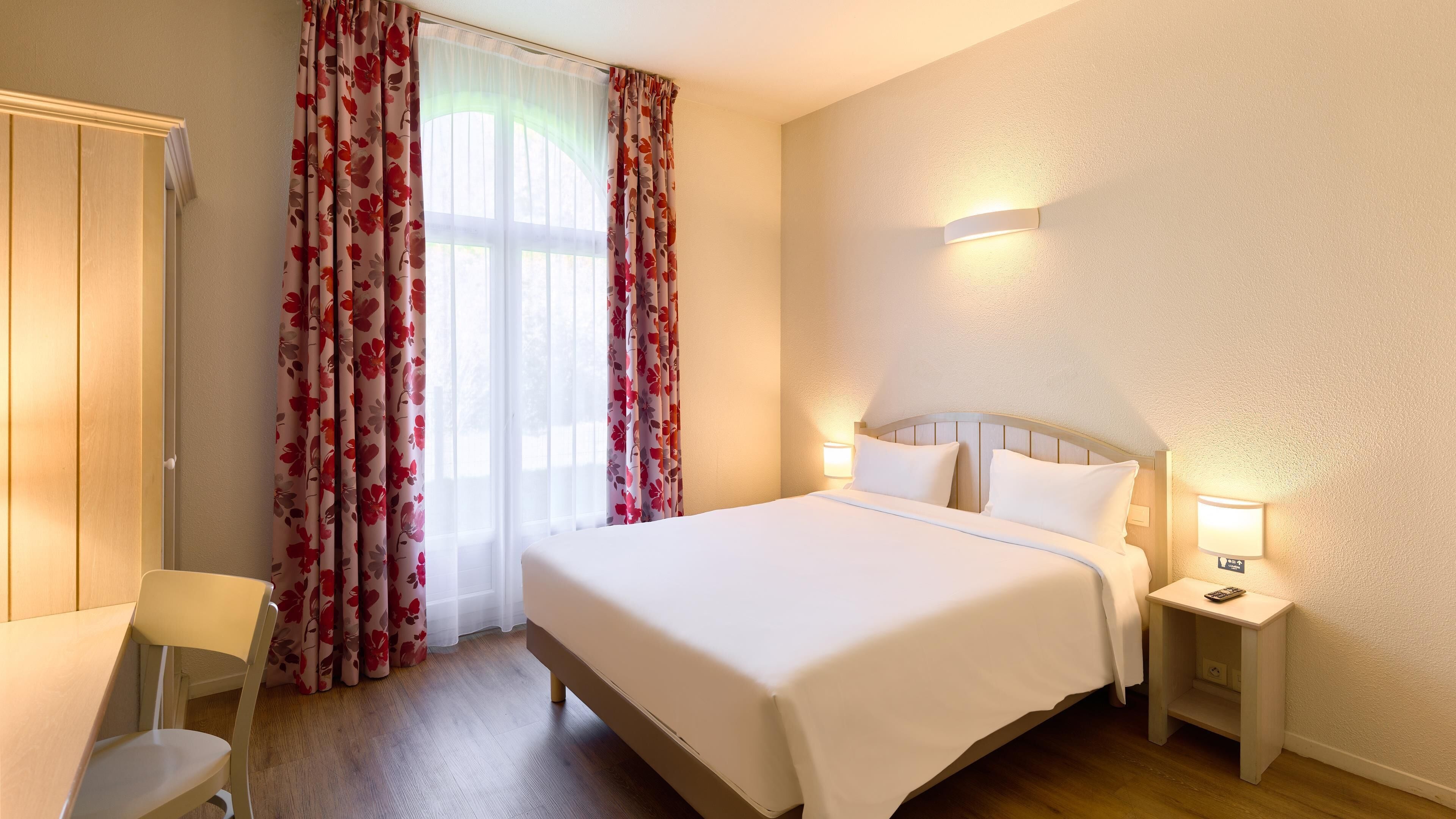 A hotel room with a bed, a chair, a nightstand with a lamp, and a window with curtains.