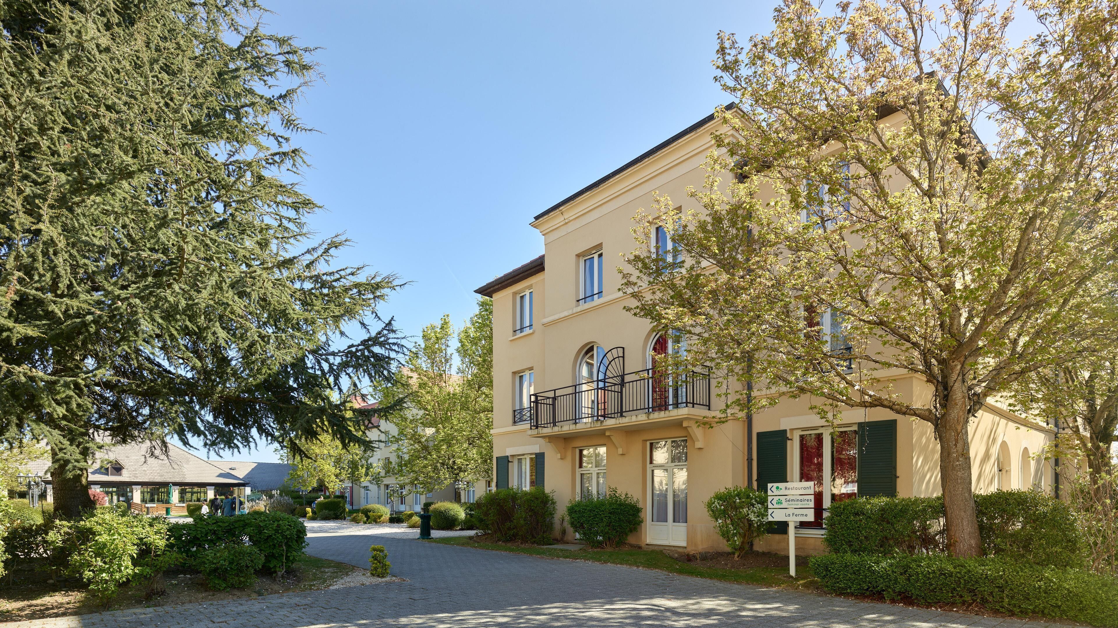 A two-story building with a balcony, surrounded by trees and a paved driveway.