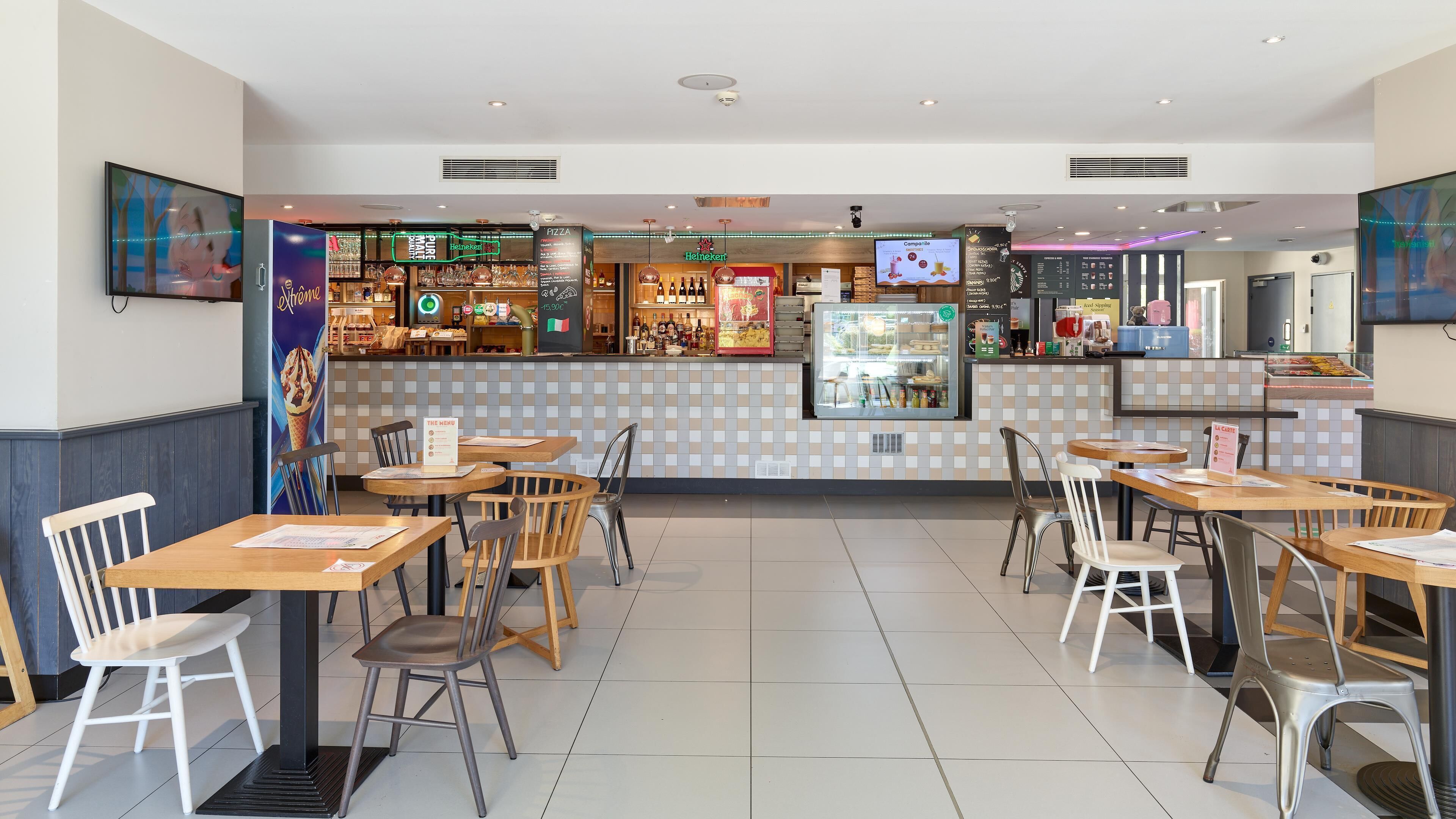 Un restaurant avec un sol carrelé, des tables et des chaises en bois, un comptoir où est affiché le menu, et une enseigne Heineken.