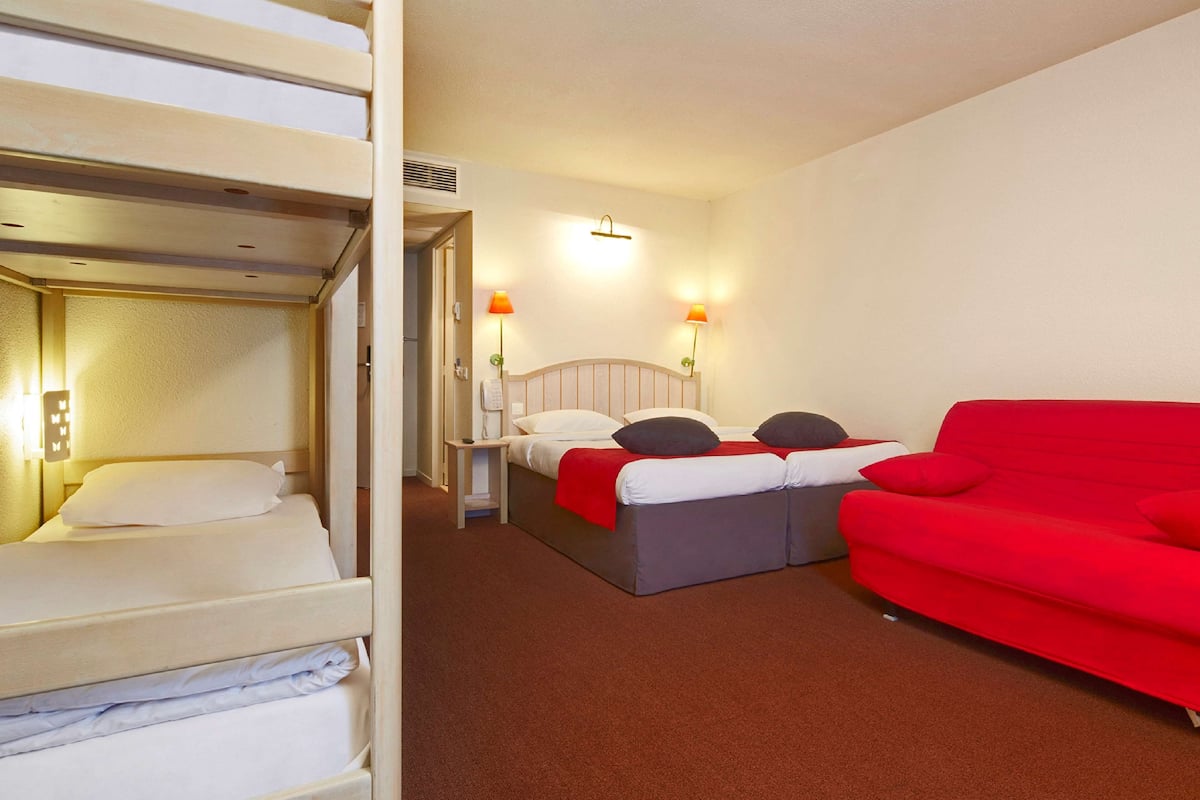 A hotel room with a bunk bed, a single bed, a red sofa, and two bedside lamps.
