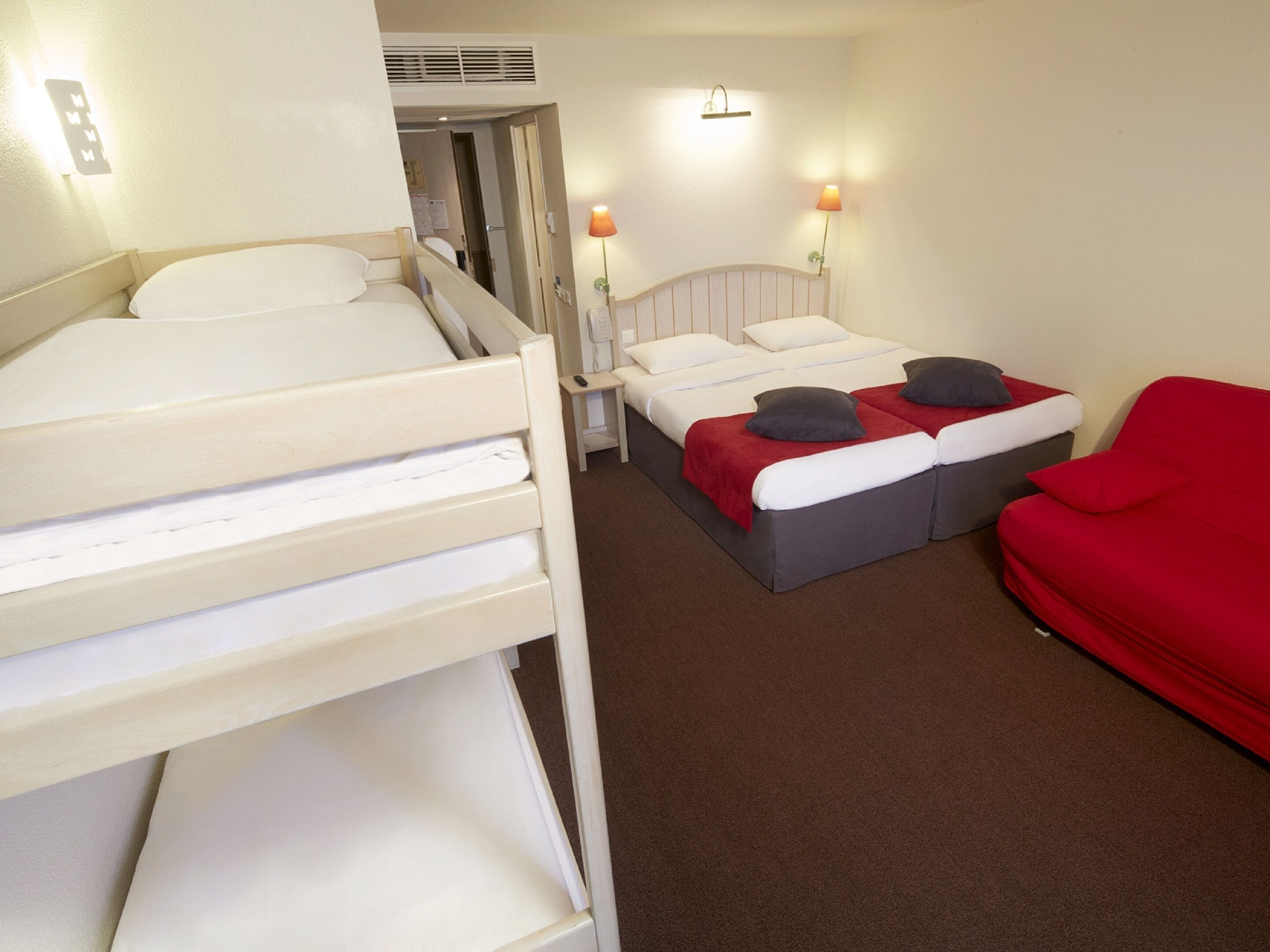 Une chambre d’hôtel avec deux lits, un canapé rouge et deux lampes fixées au mur.