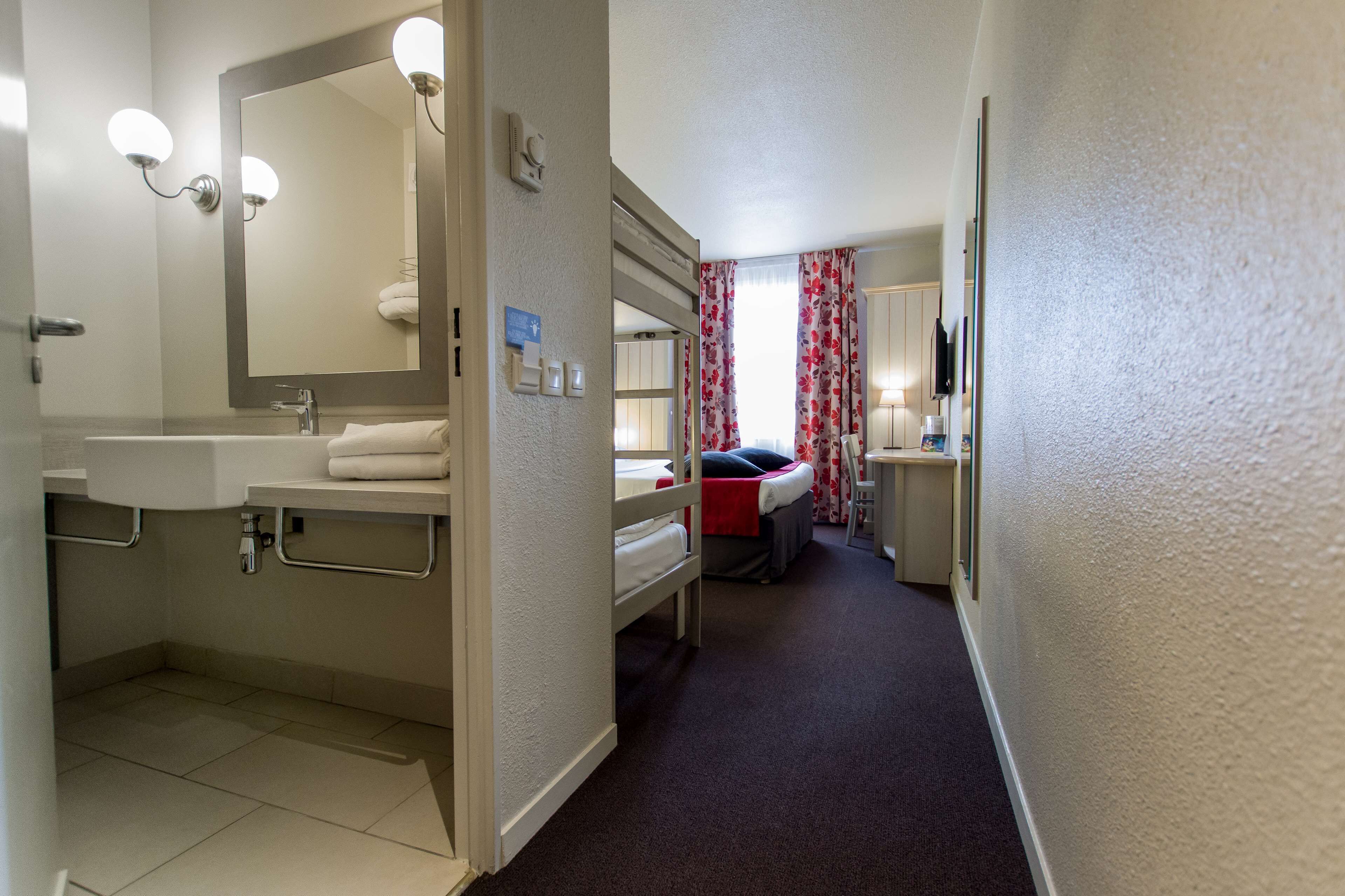 Un baño de hotel con lavamanos, espejo y perchero. Hay una cama con una manta roja y una mesita de noche con una lámpara.