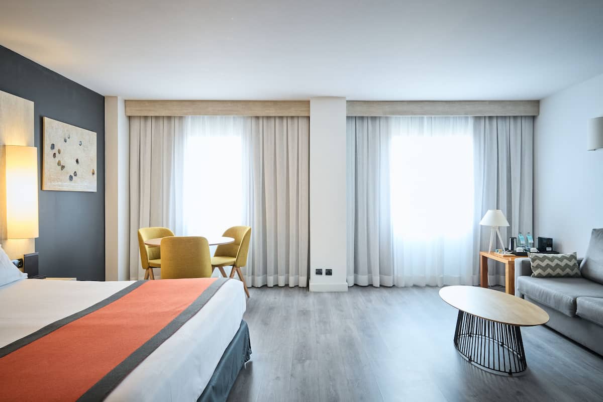 A hotel room with a bed, a round coffee table, a sofa, a desk with a lamp, and two chairs.