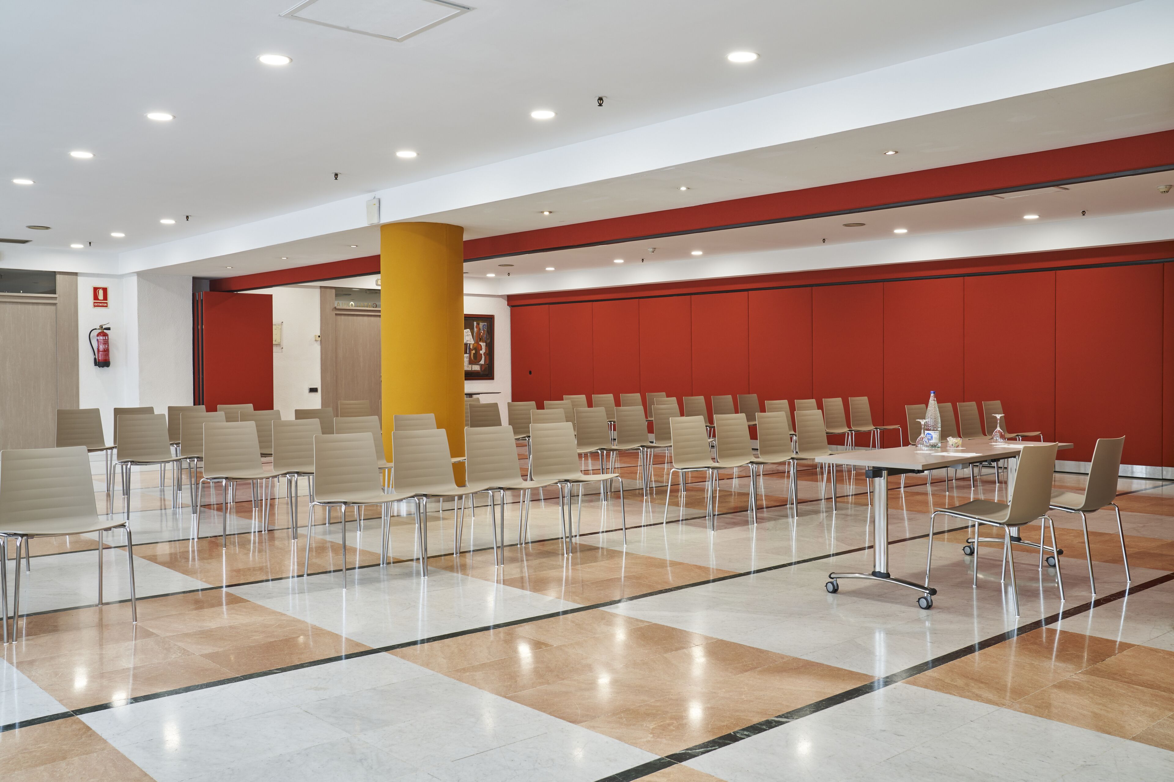 A conference room with rows of chairs and a table, white walls, and a red accent wall.