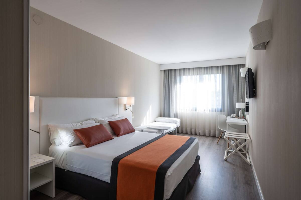 A hotel room with a large bed, bedside tables, a desk, a chair, and a TV.