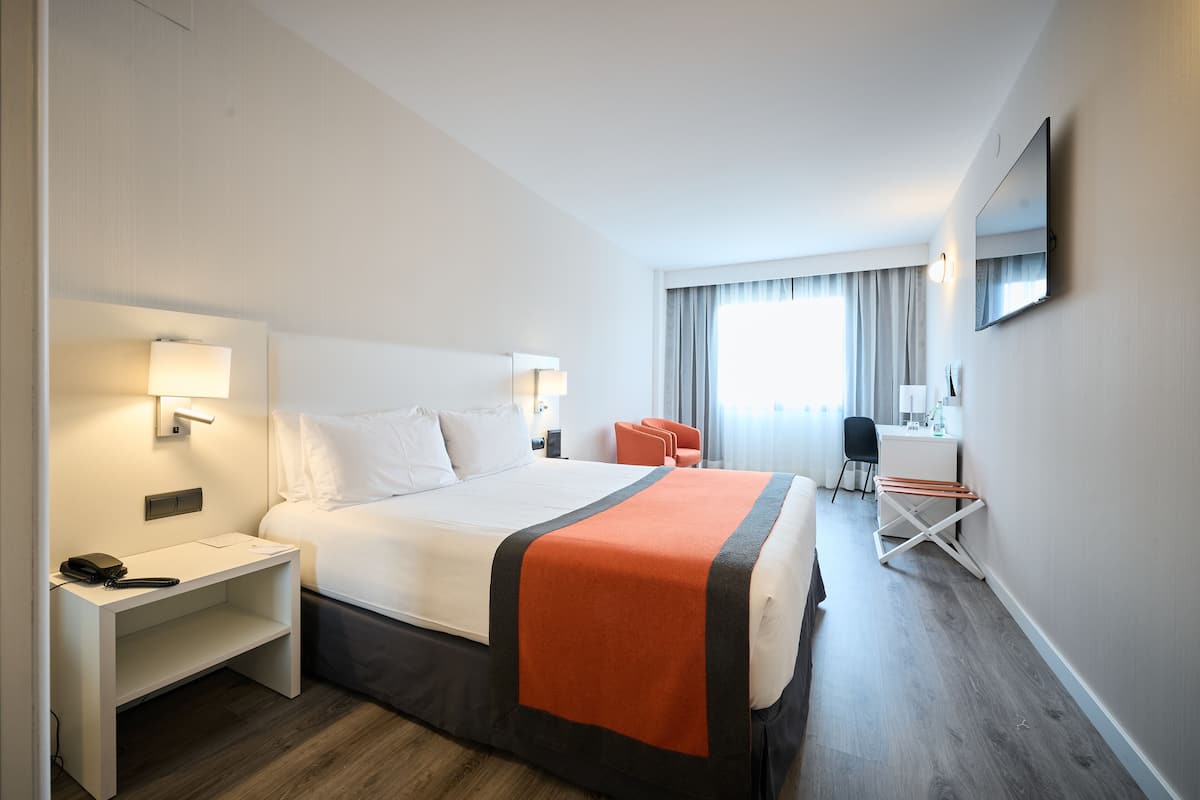 A hotel room with a large bed, a desk, a chair, and a desk lamp.