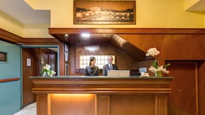 A reception desk with two staff members, a framed cityscape painting above, and a vase with white flowers. - Flower Garden Hotel Rome (Rome)