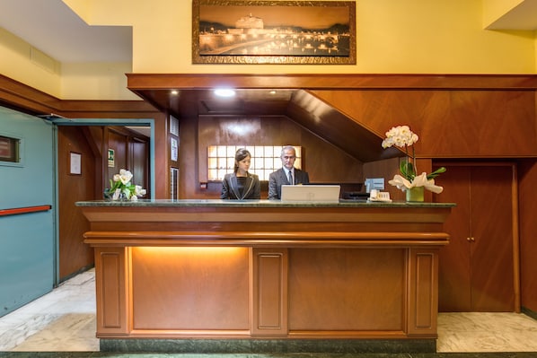 A reception desk with two staff members, a framed cityscape painting above, and a vase with white flowers. - Flower Garden Hotel Rome (Rome)