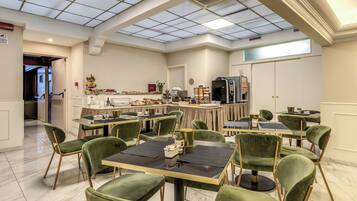 A spacious dining area with green upholstered chairs and tables, a kitchen in the background, and a high ceiling with skylights.
