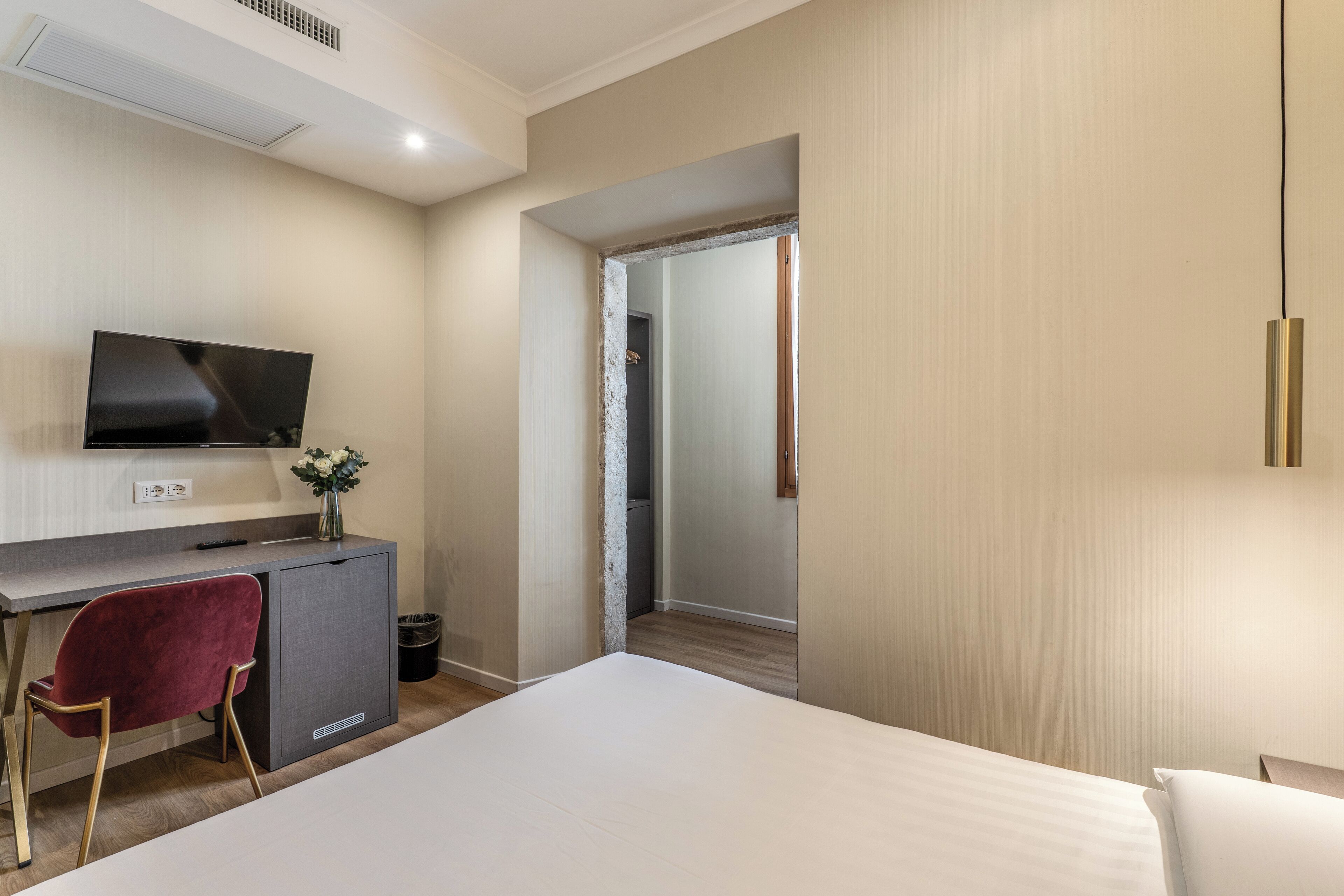 A hotel room with a bed, a desk with a TV, a red chair, and a small cabinet.