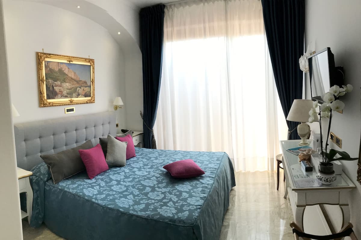 A bedroom with a tufted headboard, a bed with floral-patterned bedding, a bedside table with a lamp, a desk with a lamp, and a potted orchid.