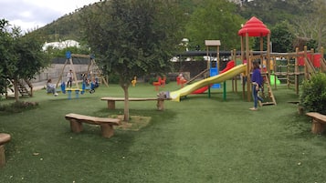 Children can play on the slide, swings, and climbing elements in this playground surrounded by lush greenery and a mountainous landscape.