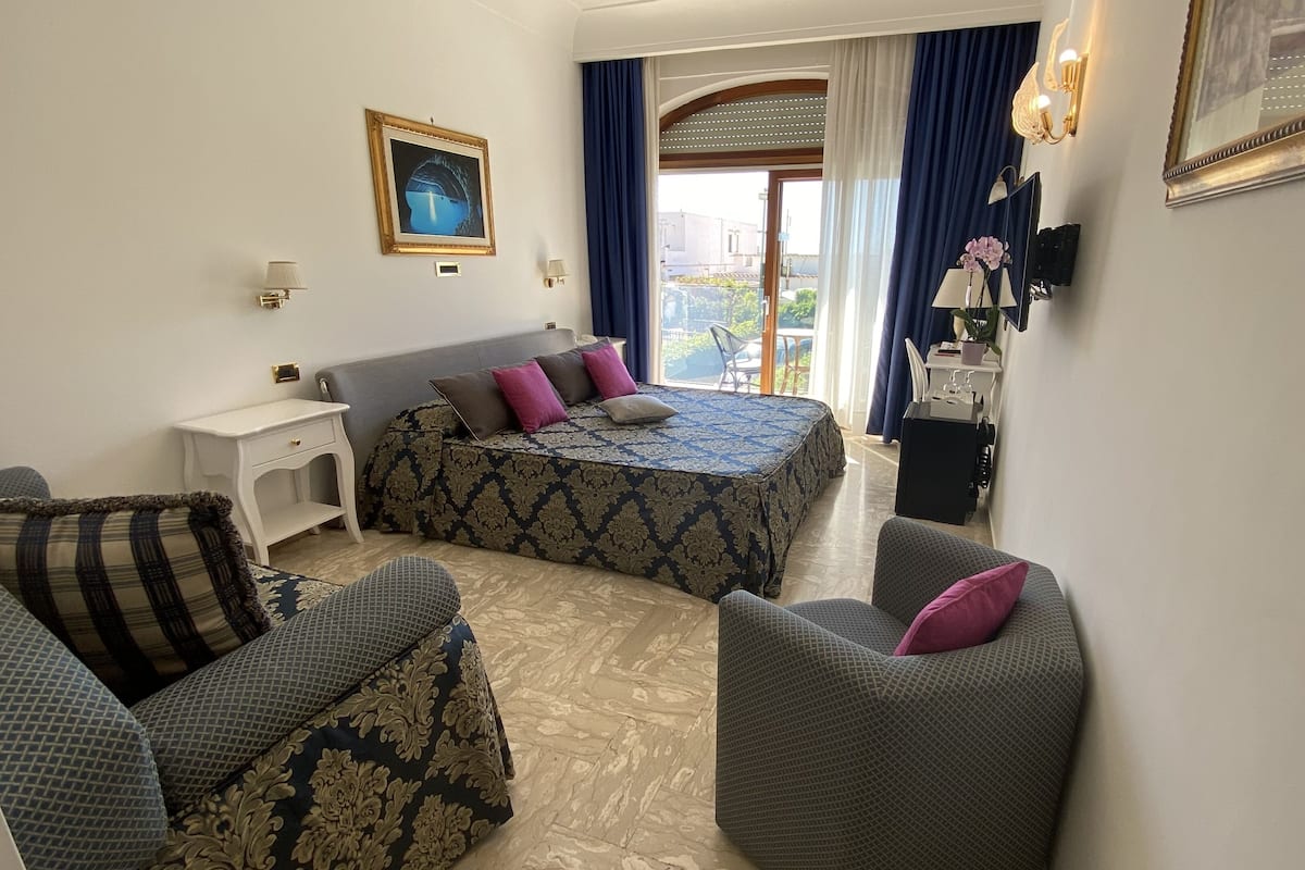 A hotel room with a bed, two armchairs, a small table, a nightstand, a window with curtains, and a wall-mounted lamp.