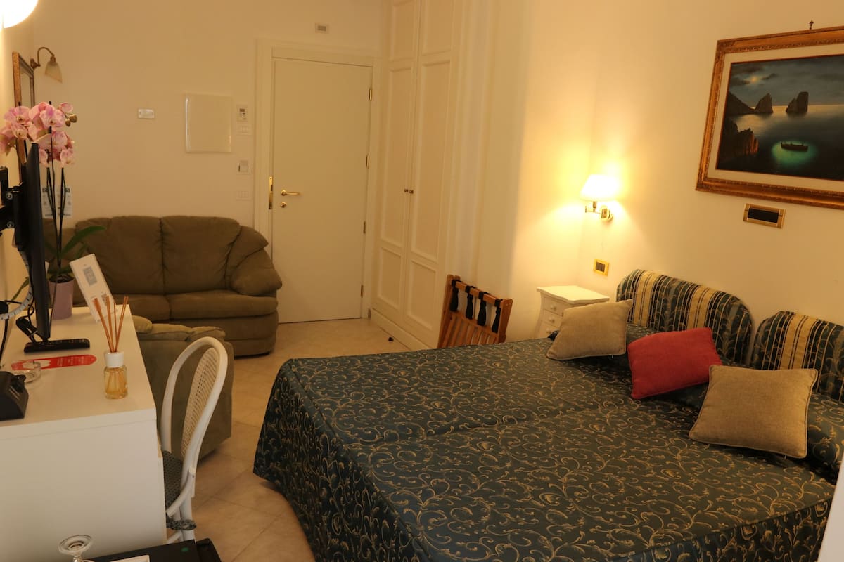 A hotel room with a bed, a sofa, a desk with a computer, a chair, and a lamp.