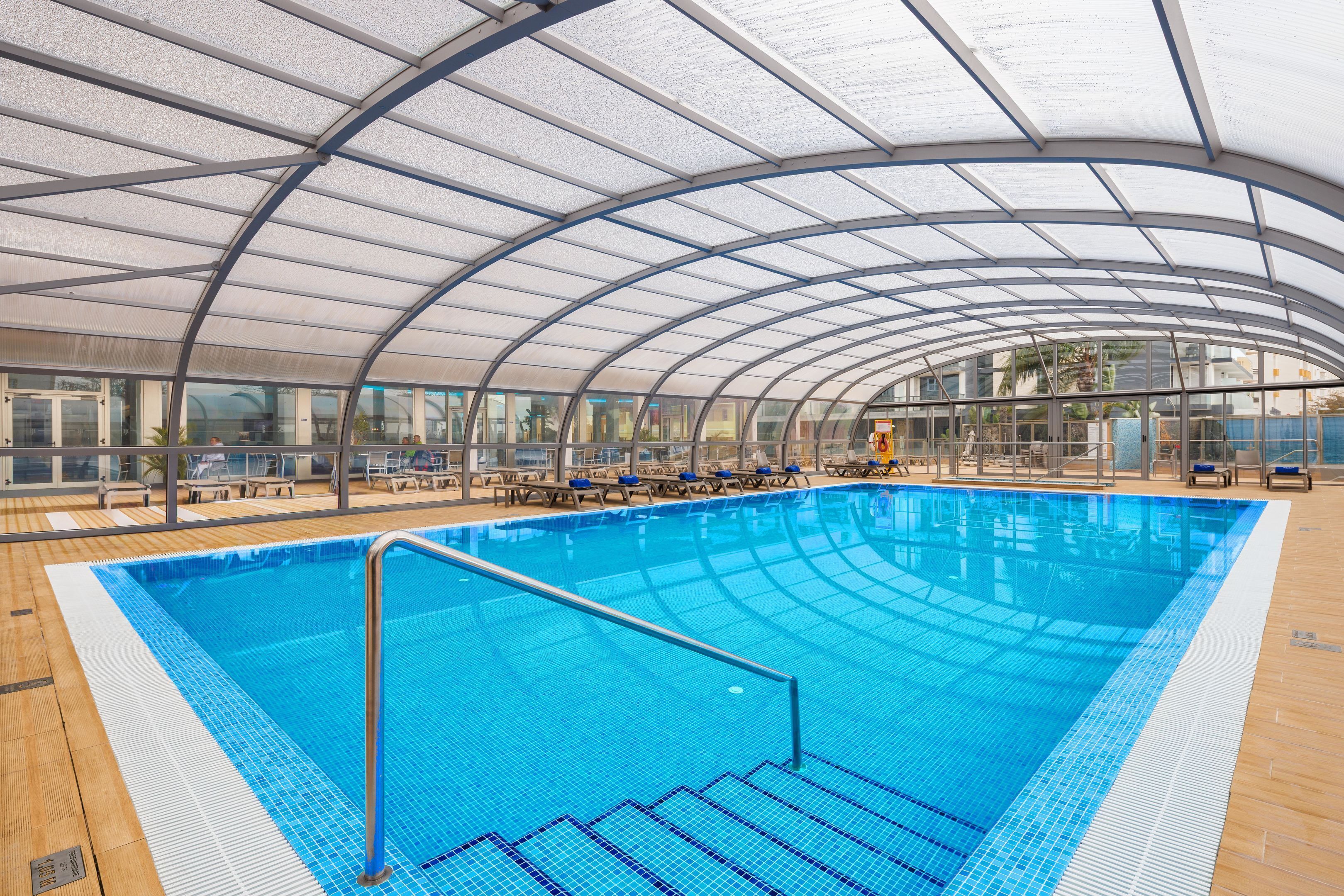 Piscina interior com teto de vidro, água cristalina e deck de madeira.