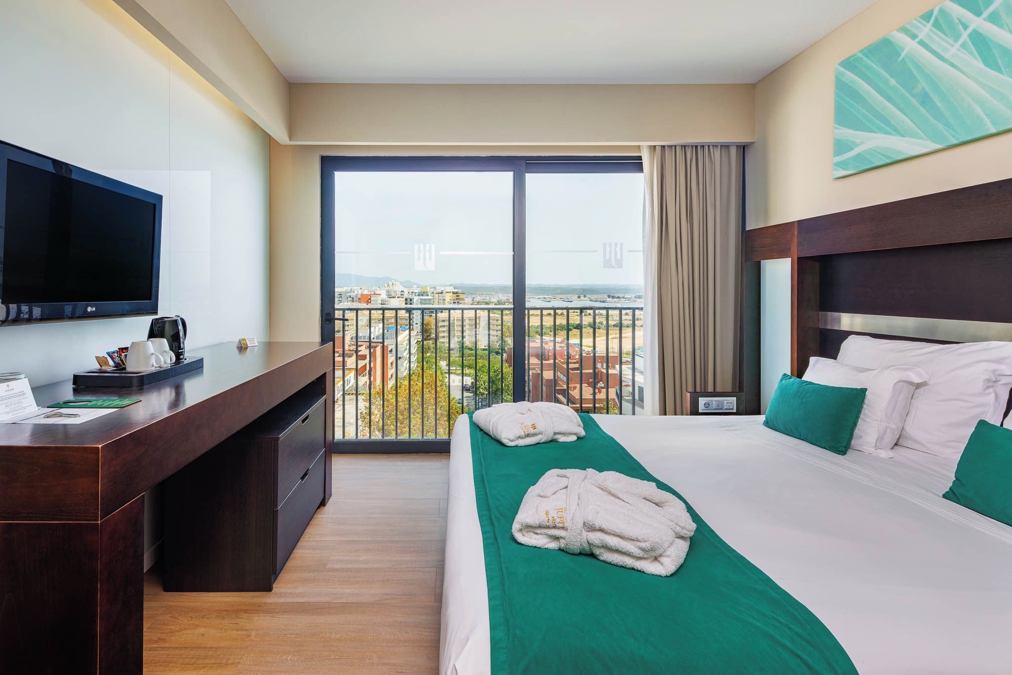 A hotel room with a bed, a desk, a television, and a balcony with a city view.
