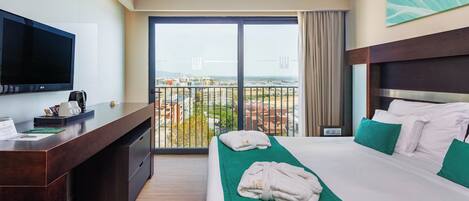 A hotel room with a bed, a desk, a television, and a balcony with a city view.