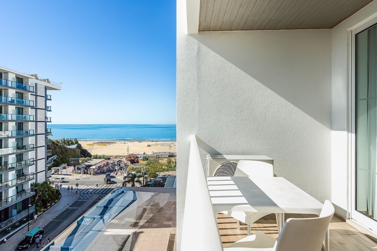 A balcony with a table and chairs, overlooking a beach and a modern building with balconies.