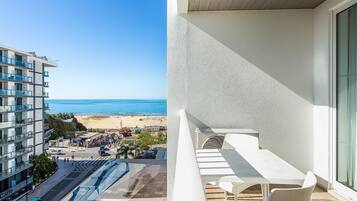 Varanda com mesa e cadeiras, vista para uma praia e um edifício moderno com varandas.