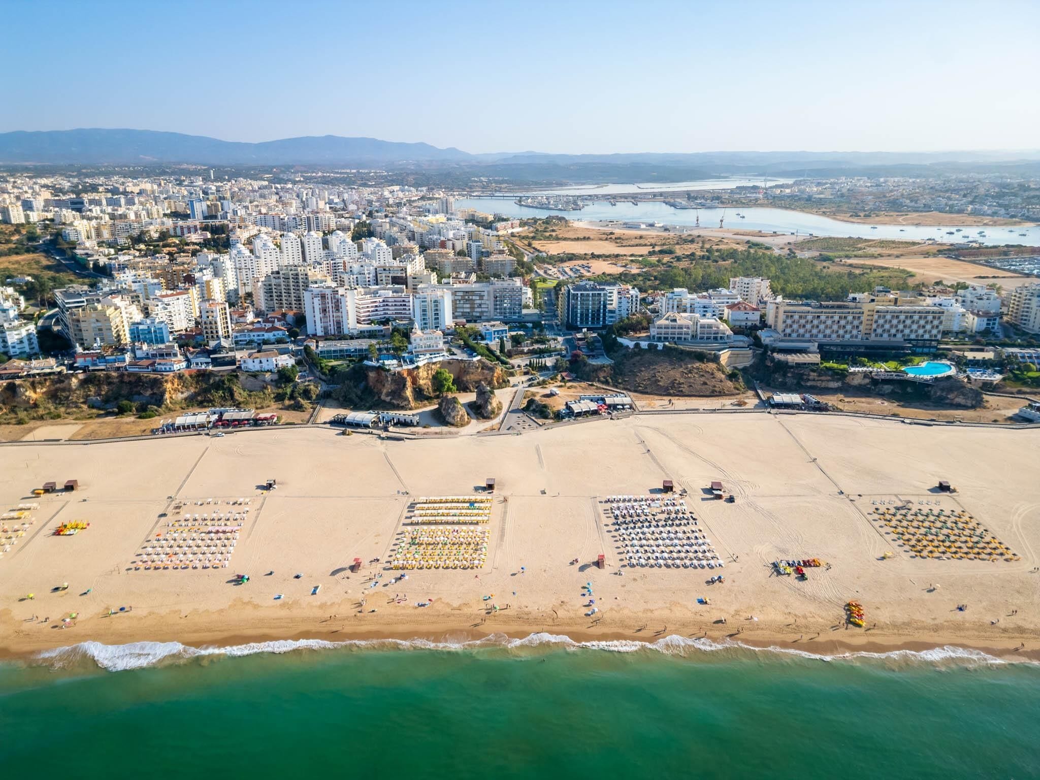 An aerial view of a coastal city with a wide beach, numerous buildings, and a clear view of the sea.