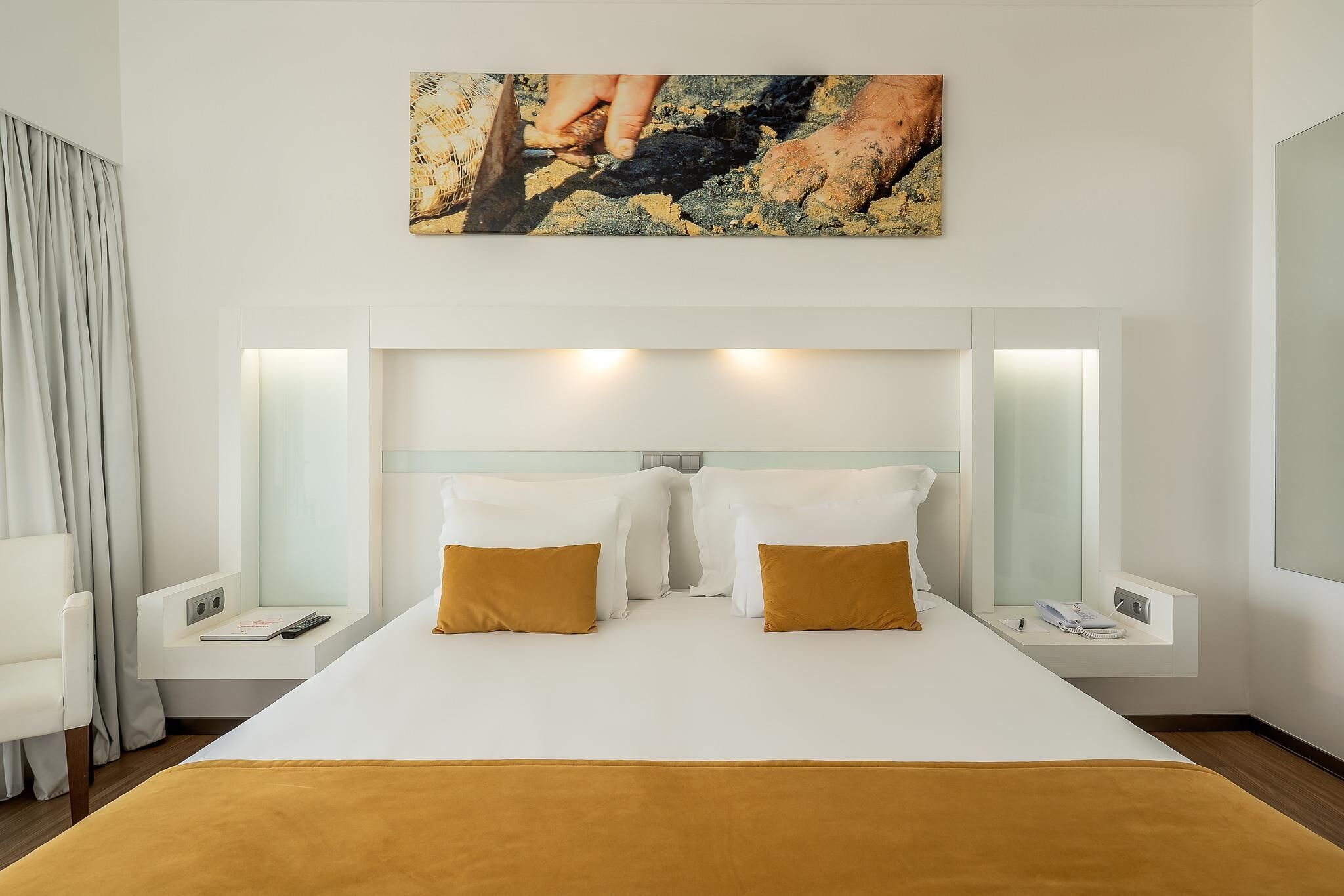 A hotel room with a large bed, two bedside tables, a nightstand, a painting above the bed, and a window with curtains.