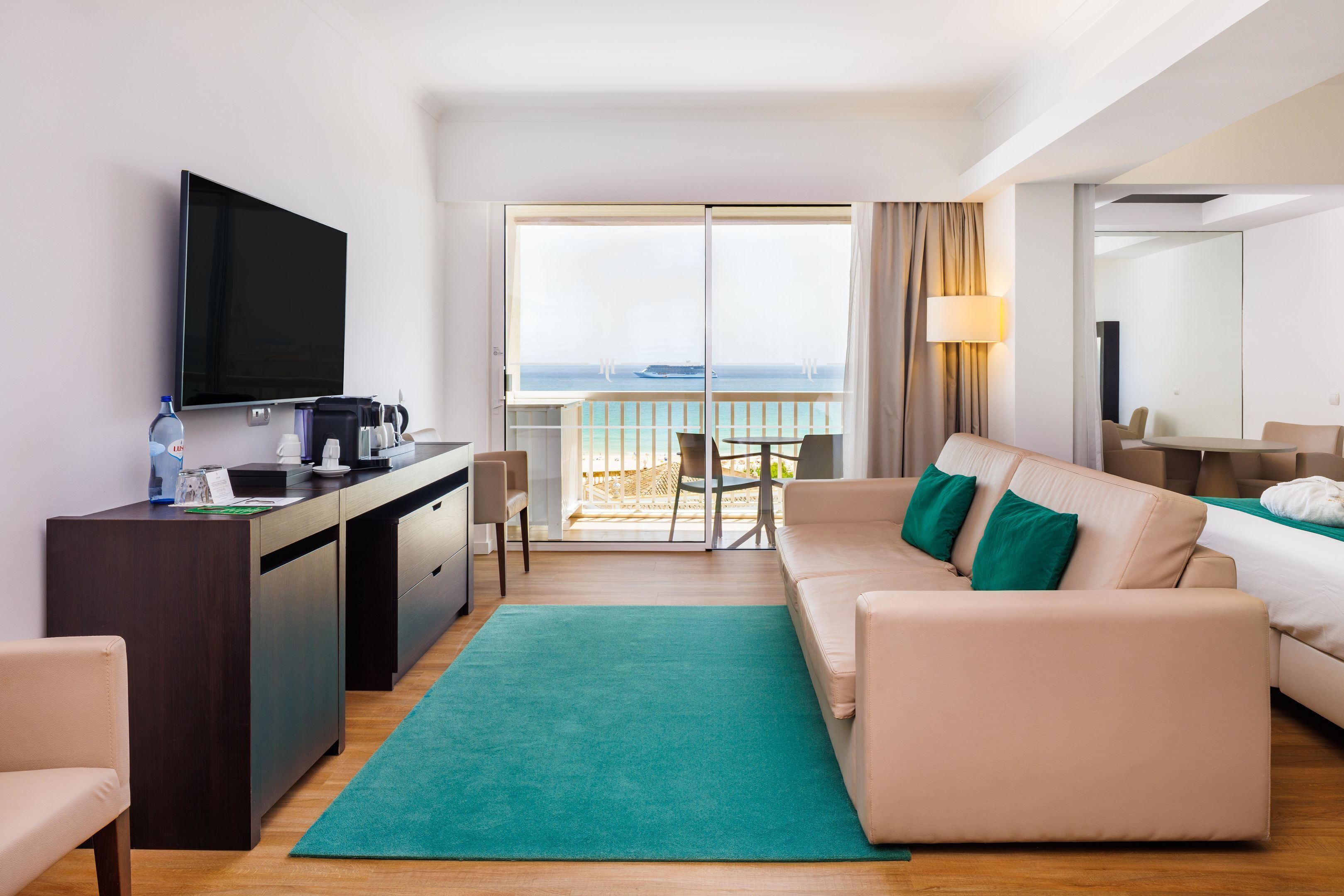A modern hotel room with a balcony, a sofa, a TV, and a desk with a coffee maker.