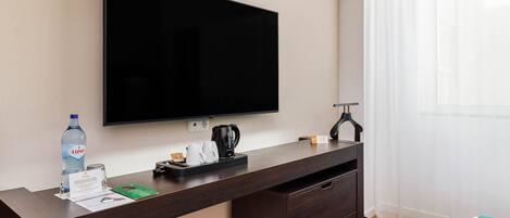 Habitación de hotel con un escritorio de madera oscura, un televisor de pantalla plana, una botella de agua, una cafetera y una cama con manta verde.